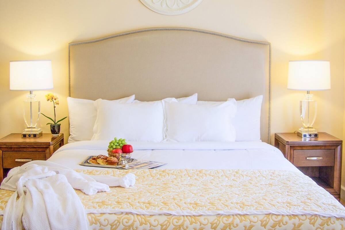 A hotel room with a large bed, two bedside tables with lamps, a decorative wall piece, and a fruit bowl on the bed.