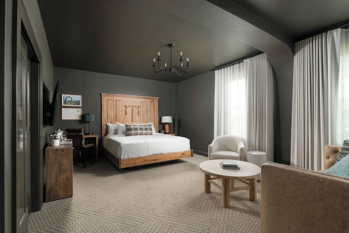 A modern hotel room with a large bed, a wooden nightstand, a desk, a chair, a round coffee table, and a chandelier.