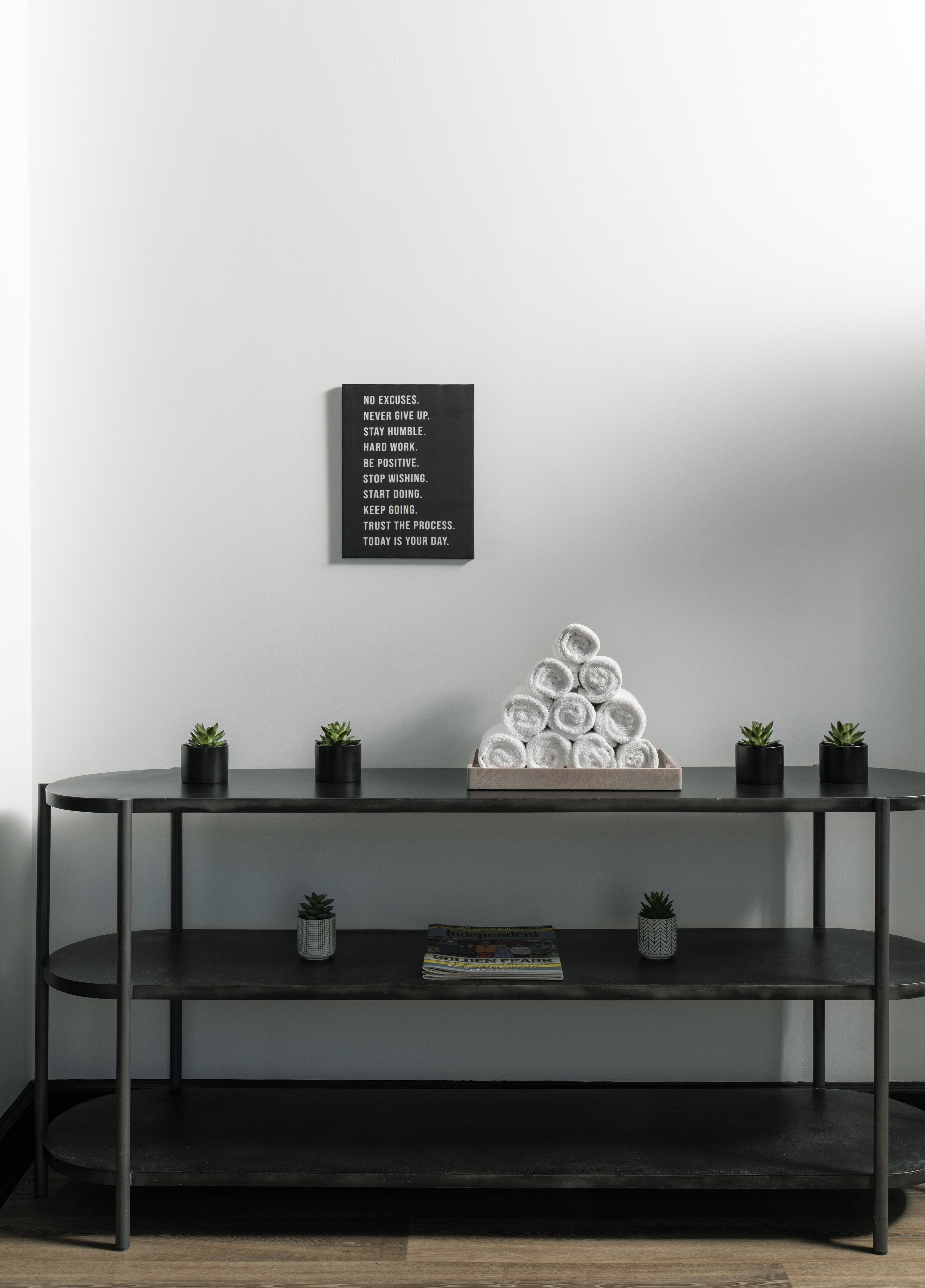 A minimalist room with a dark metal console table, a wall-mounted motivational quote, and a stack of rolled towels.