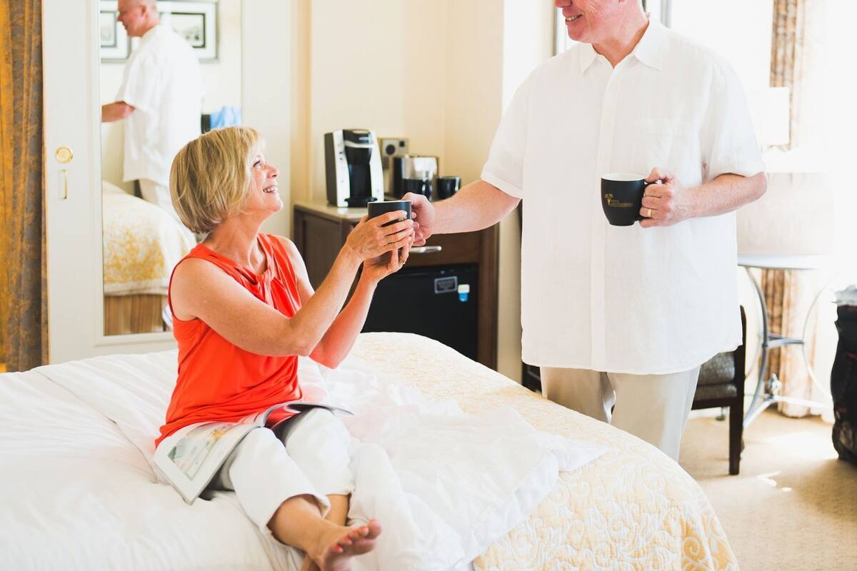A hotel room with a bed, a bedside table with a coffee maker, and a mirror reflecting a person in the background.