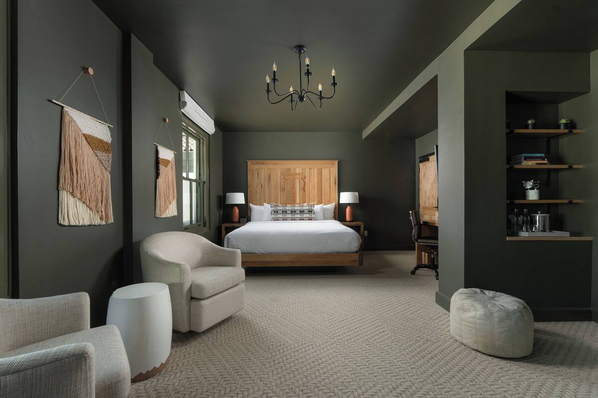 A bedroom with a large bed, two bedside tables with lamps, a chair, a round ottoman, and a wooden wardrobe.