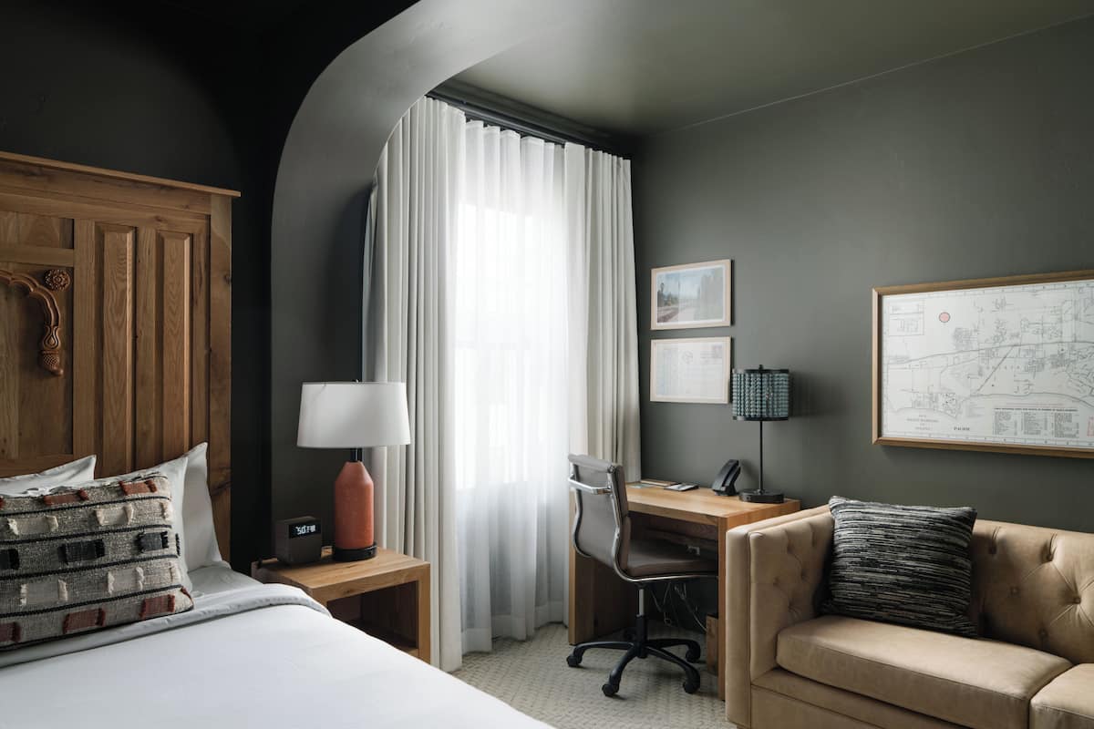 A hotel room with a bed, a desk, a chair, a sofa, a lamp, and curtains.