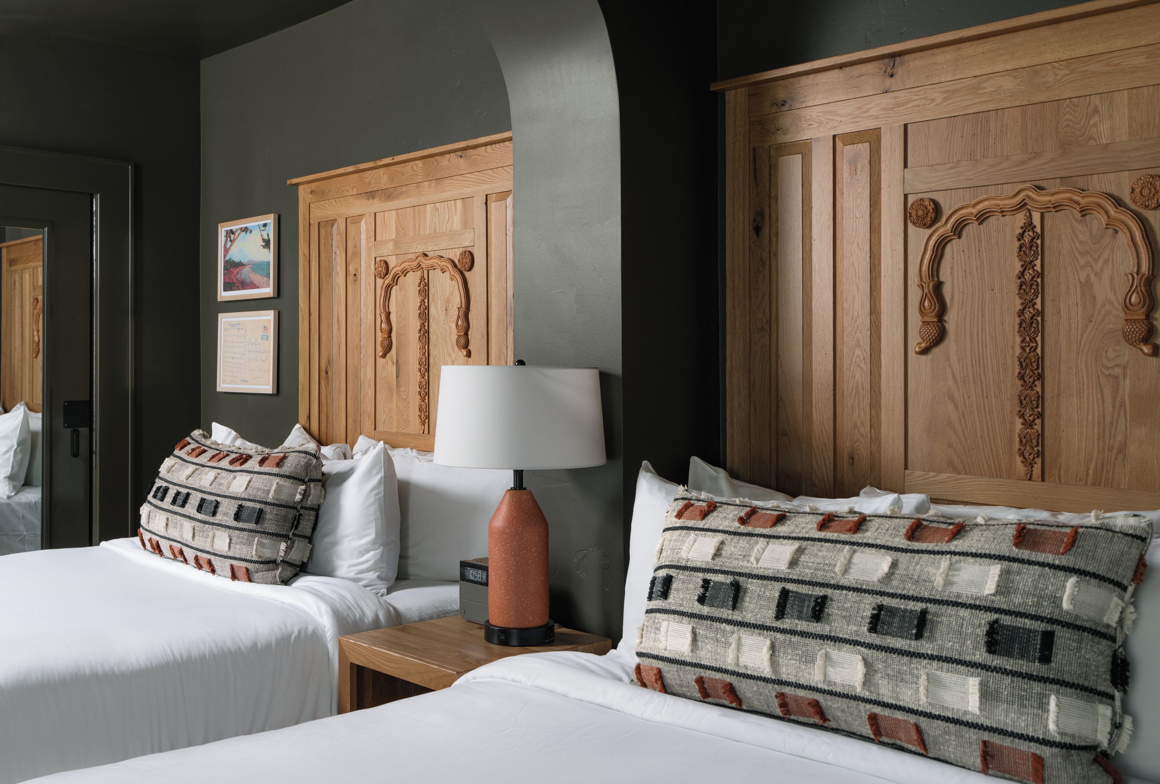 Habitación de hotel con dos camas, cabecera de madera, mesita de noche con lámpara y un cuadro en la pared.