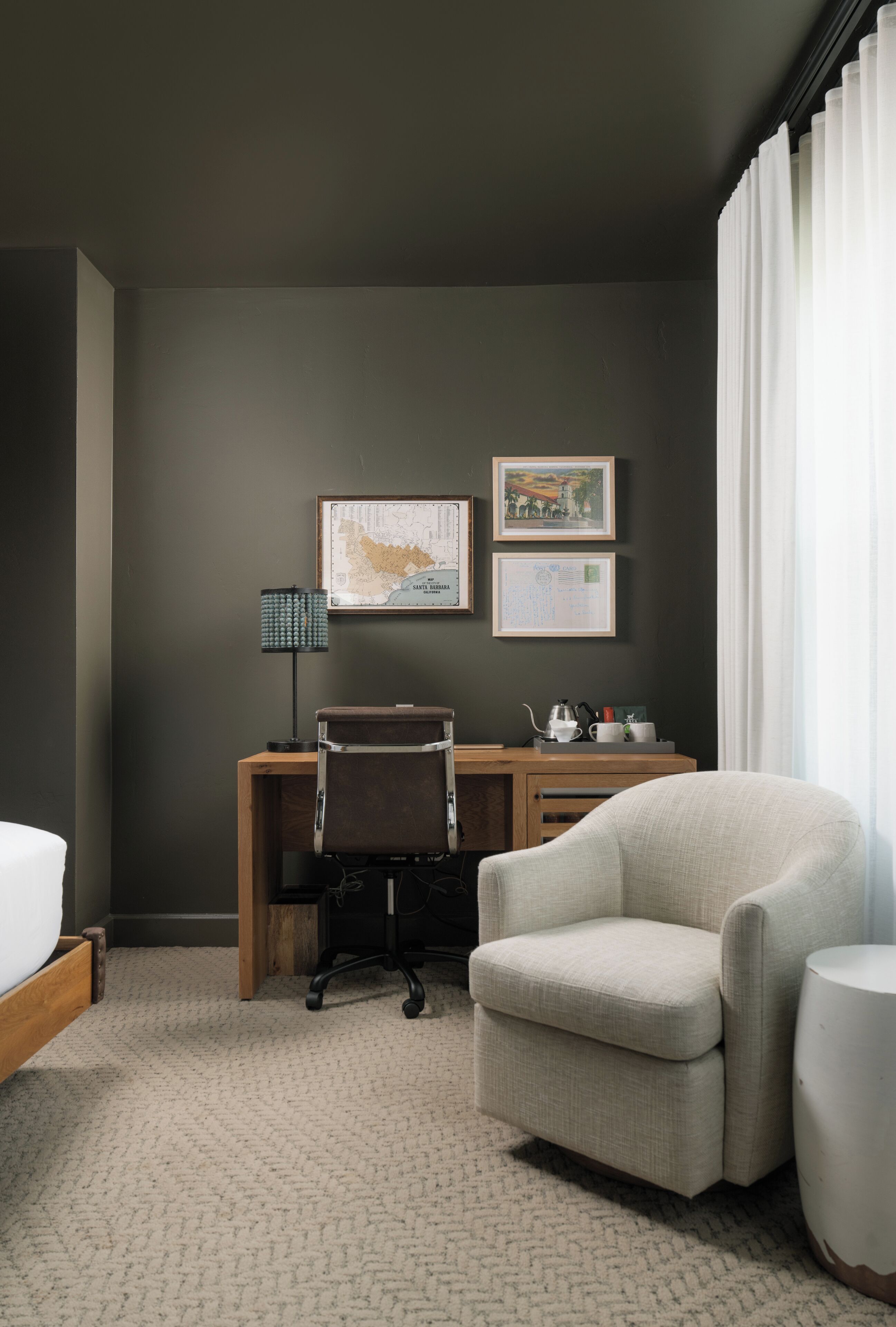 A hotel room with a bed, a desk, a chair, a lamp, and two framed pictures on the wall.