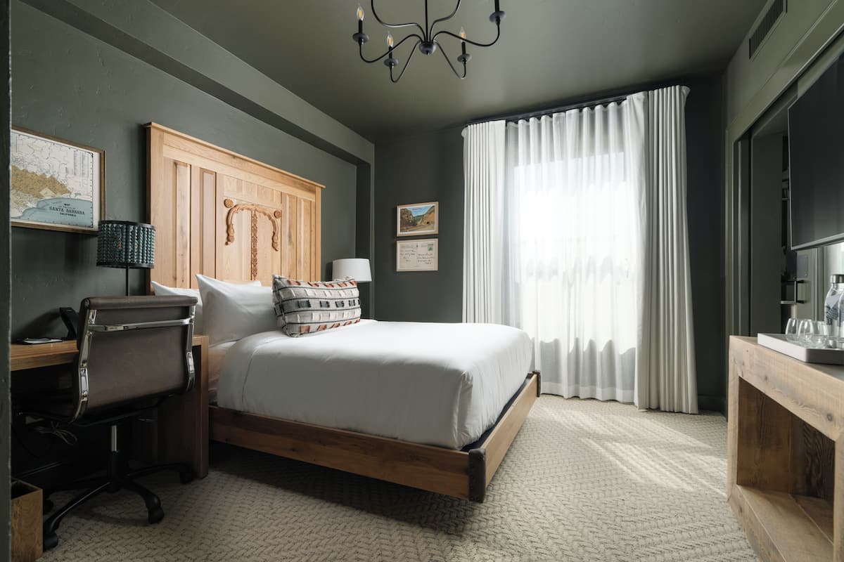 A bedroom with a wooden bed, a desk with a chair, a lamp, and a large window with curtains.