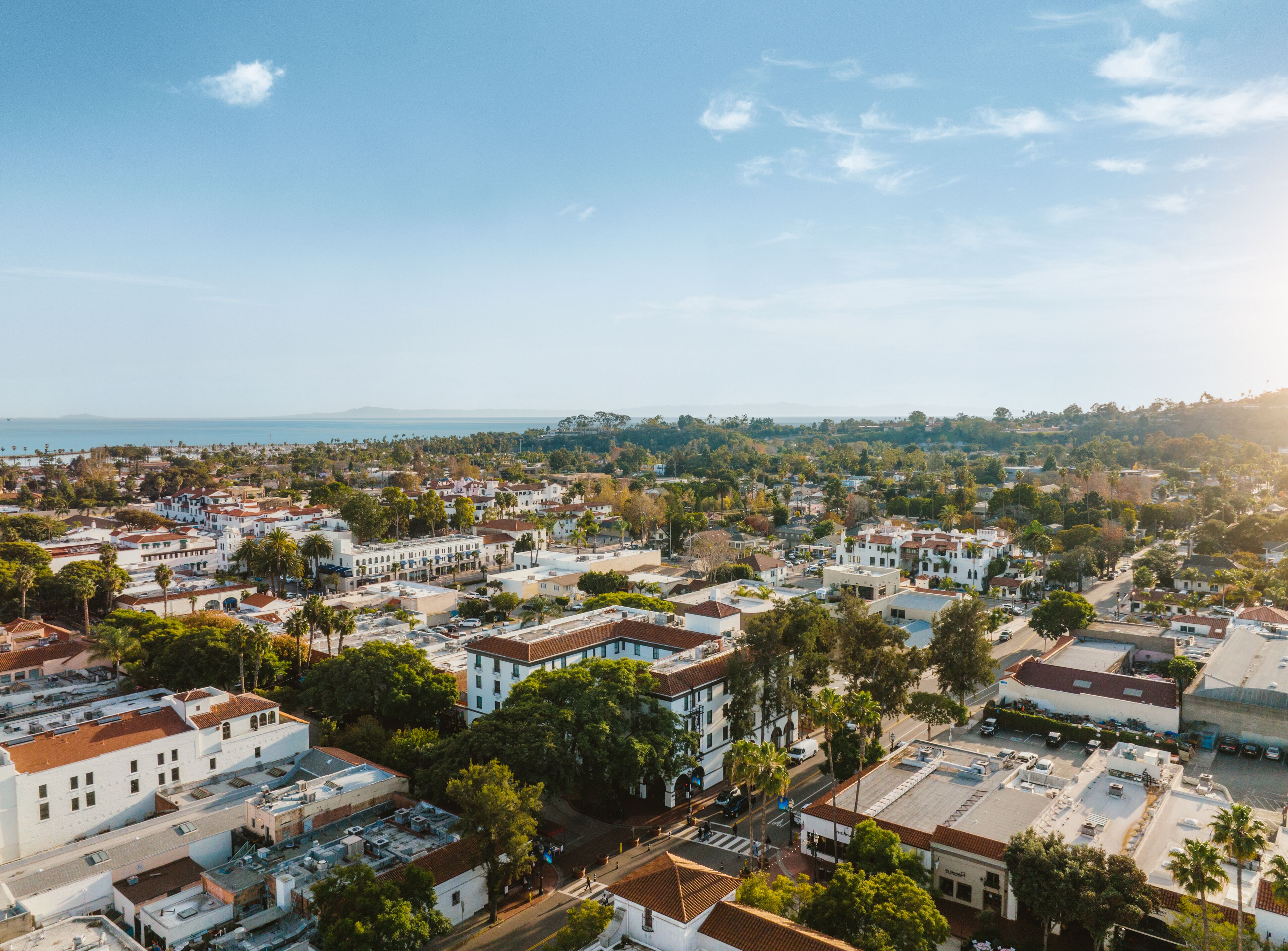 An aerial view of a coastal town with white buildings, green trees, and a clear sky.