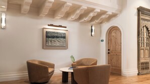 A room with a wooden floor, two brown armchairs, a round table with a book and a plant, a framed painting on the wall, and a wooden door with intricate carvings.
