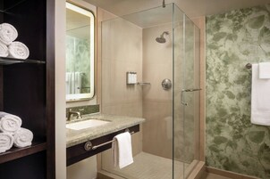 Un baño moderno con ducha de vidrio, lavabo con encimera de mármol y espejo superior. Hay toallas en un estante y otra colgando en la pared.