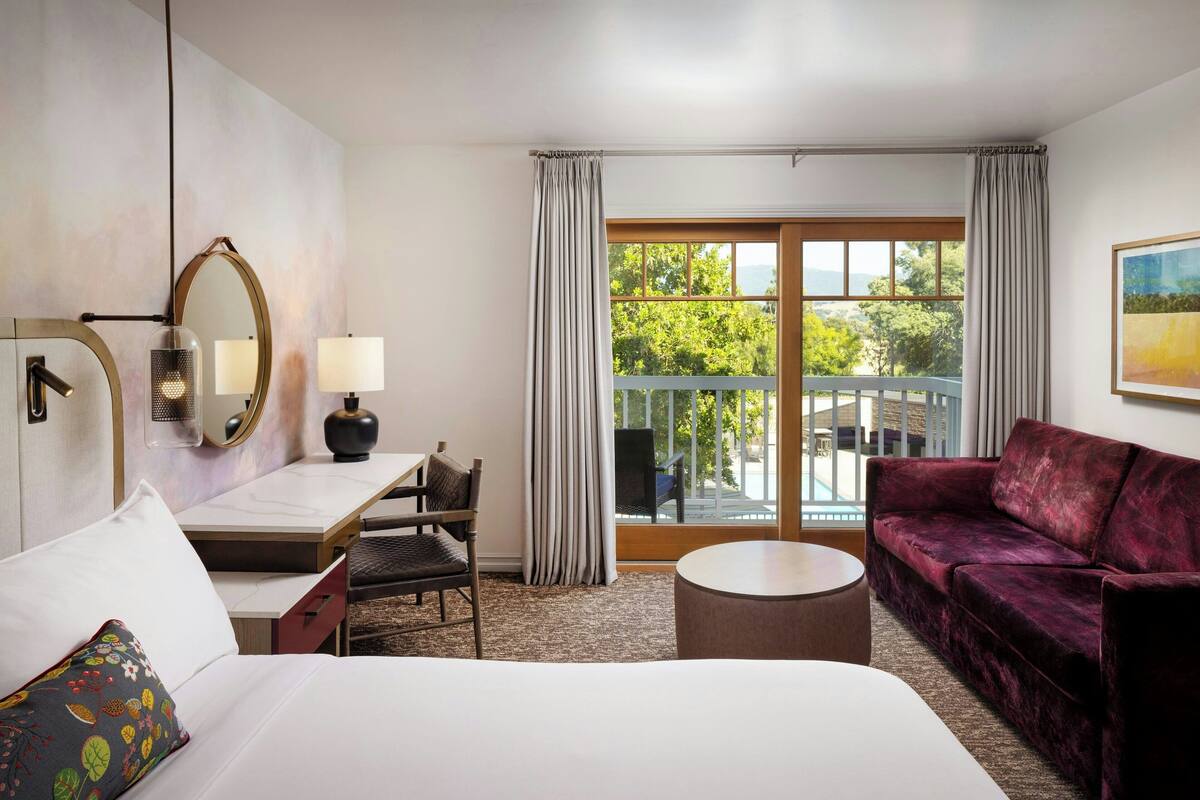 A hotel room with a bed, a desk with a lamp, a sofa, a round table, and a large window with curtains.
