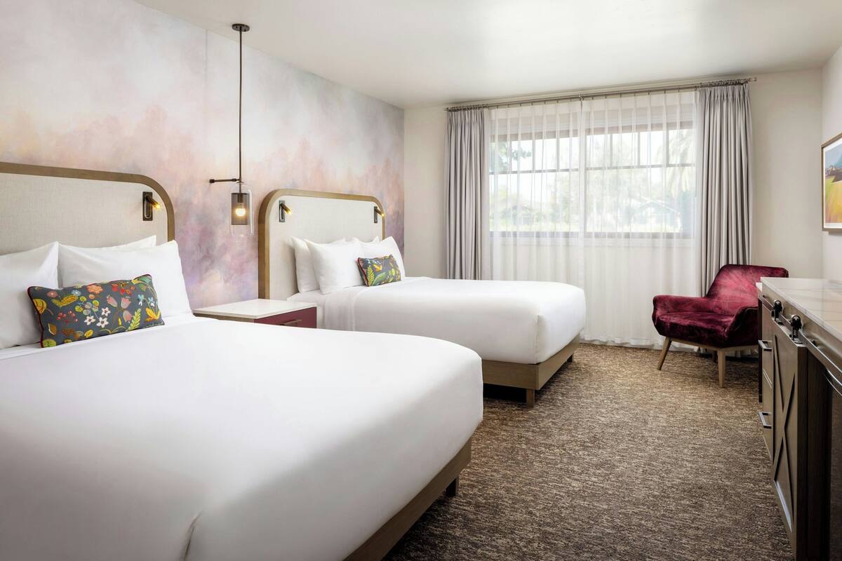A hotel room with two beds, a desk, a chair, and a large window with curtains.