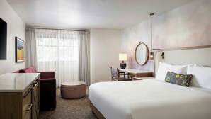 A hotel room with a large bed, a desk, a chair, a round mirror, a lamp, and a window with curtains.