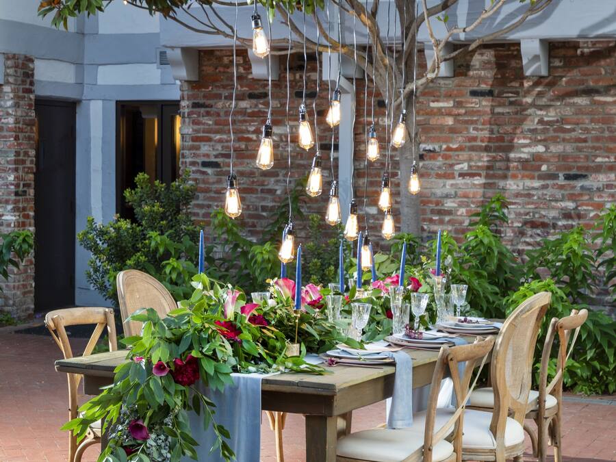 An outdoor dining area with a brick wall, wooden chairs, and a table set with flowers and candles.