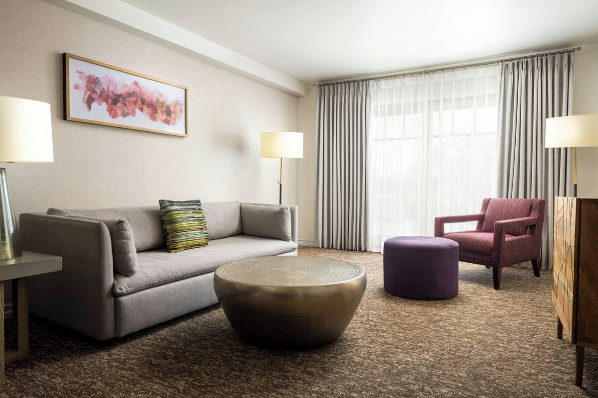 A hotel room with a grey sofa, a round coffee table, a purple ottoman, a pink armchair, and a large window with curtains.