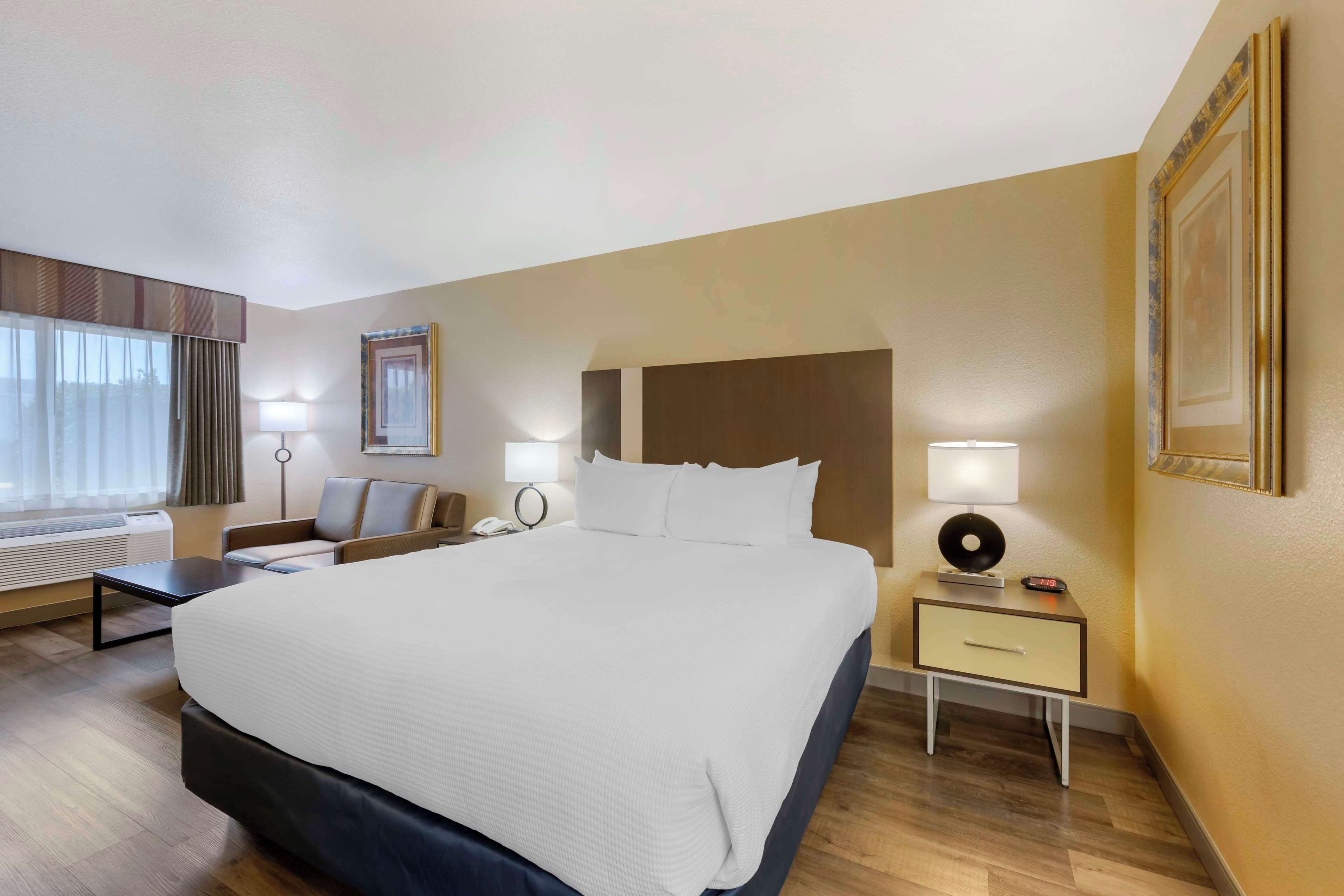 Una habitación de hotel con una cama grande, una mesita de noche con lámpara, una silla, un escritorio y un ventanal con cortinas.