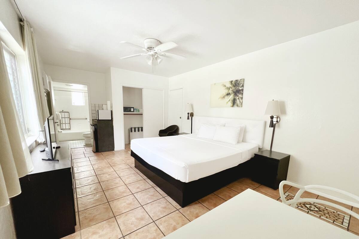 A hotel room with a bed, a desk, a chair, a ceiling fan, a bathroom, and a small kitchen area.