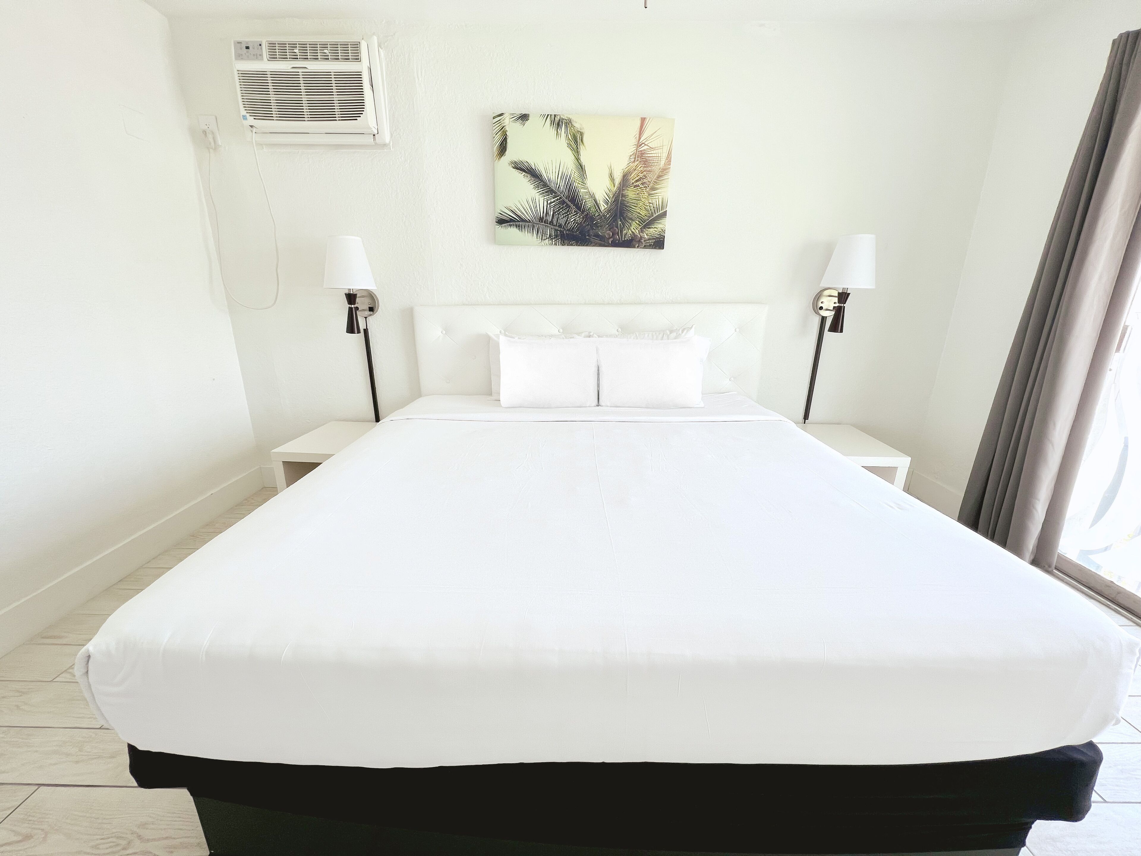 Un grand lit avec du linge de lit blanc, deux tables de chevet, un climatiseur fixé au mur et un tableau sur le thème des pays tropicaux.