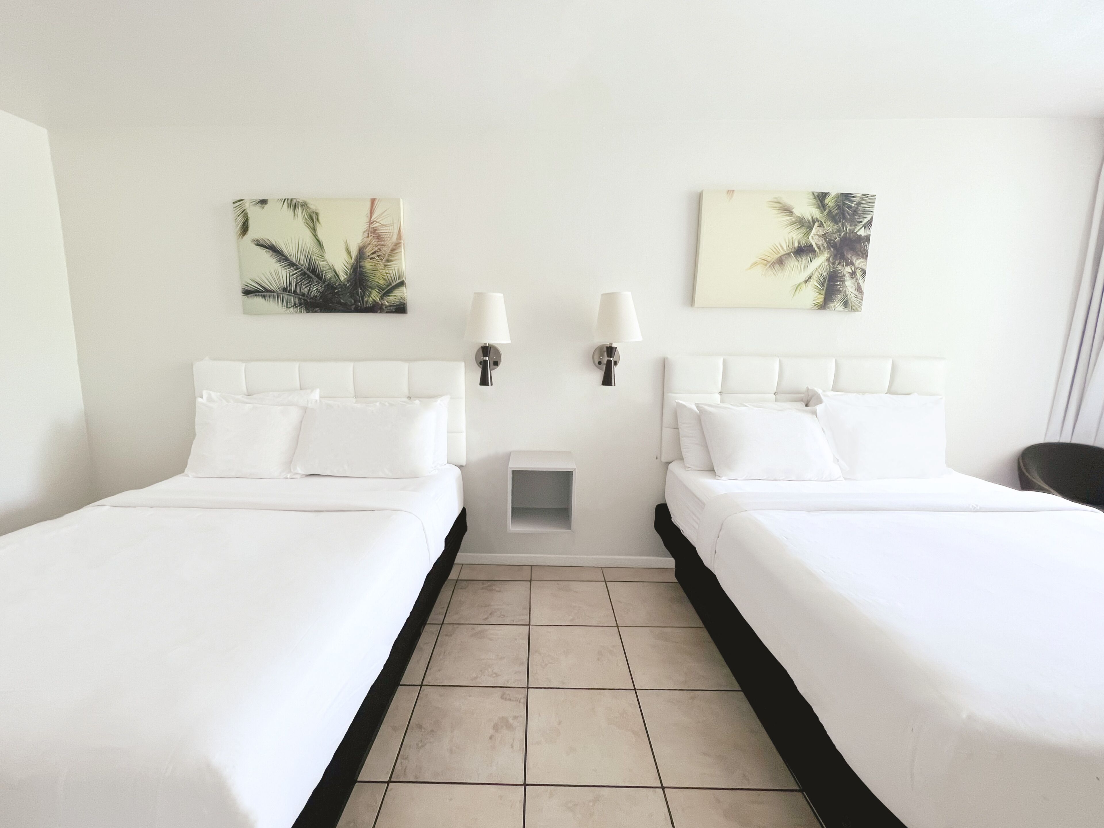 Una habitación de hotel con dos camas, ropa de cama blanca y dos lámparas fijadas a la pared.