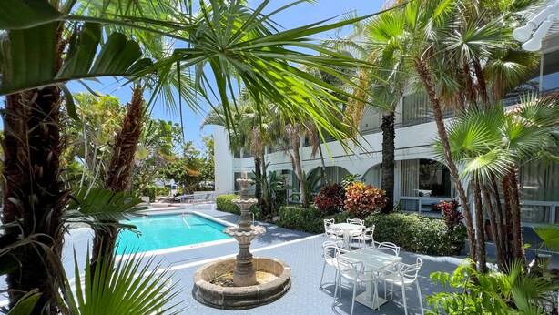 A pool area with a fountain, surrounded by palm trees and a seating area with white tables and chairs.