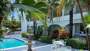 A pool area with a stone fountain, white outdoor seating, and lush tropical plants.