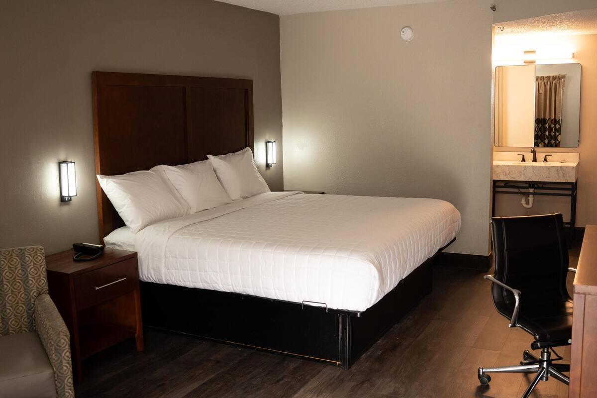 A hotel room with a bed, bedside table, chair, and a bathroom with a mirror and sink.