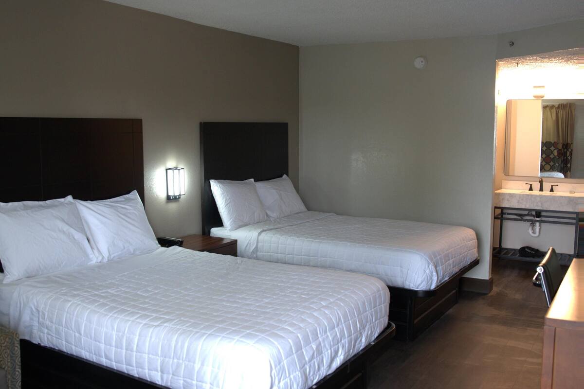 A hotel room with two beds, a nightstand, a lamp, a mirror, and a bathroom with a sink and mirror.