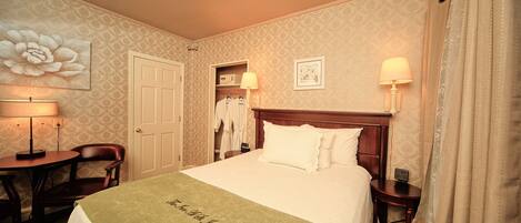 A hotel room with a neatly made bed, a bedside table with a lamp, a chair, a wardrobe, and a large floral artwork on the wall.