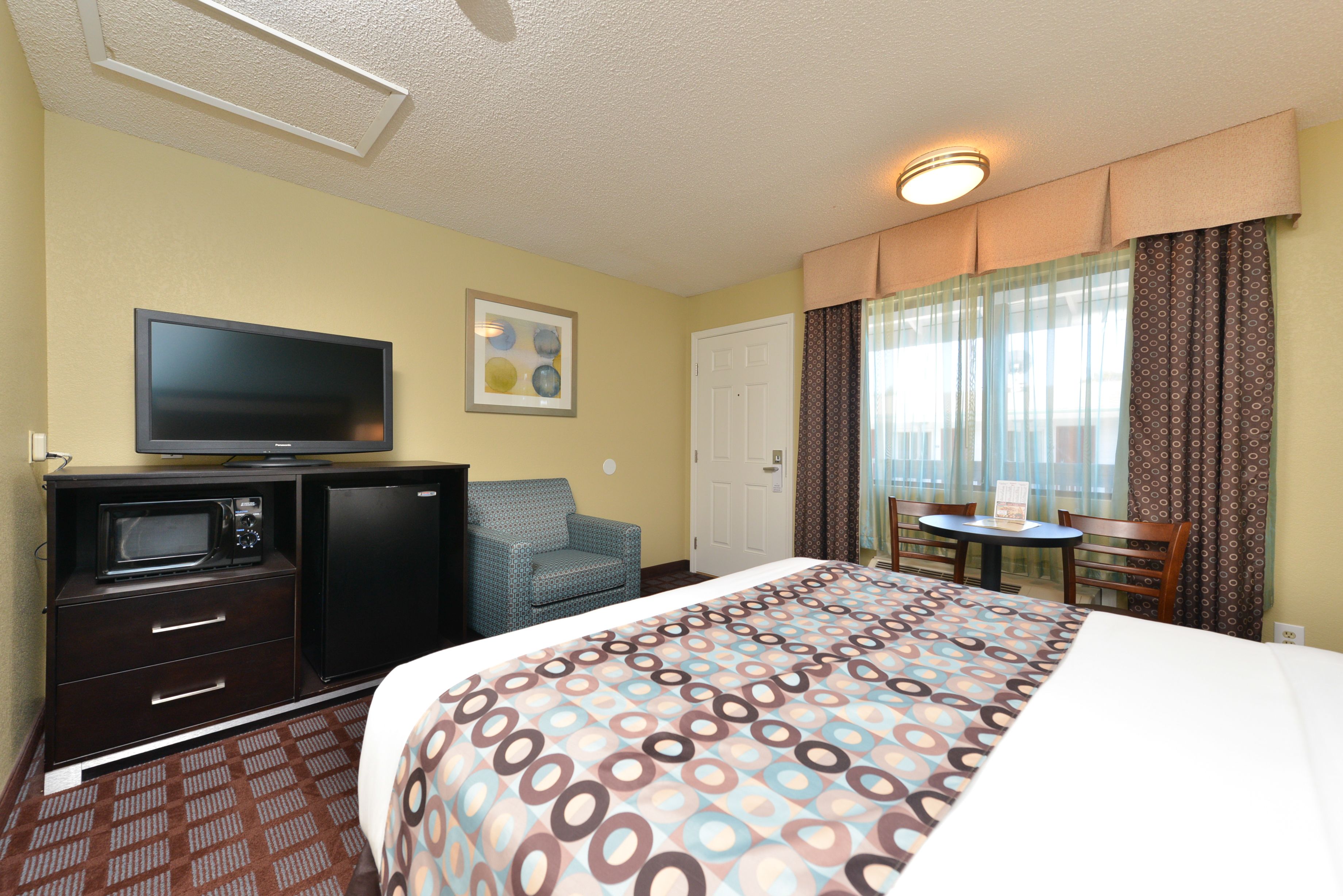 A hotel room with a bed, a TV, a desk with a microwave, a chair, and a small table.