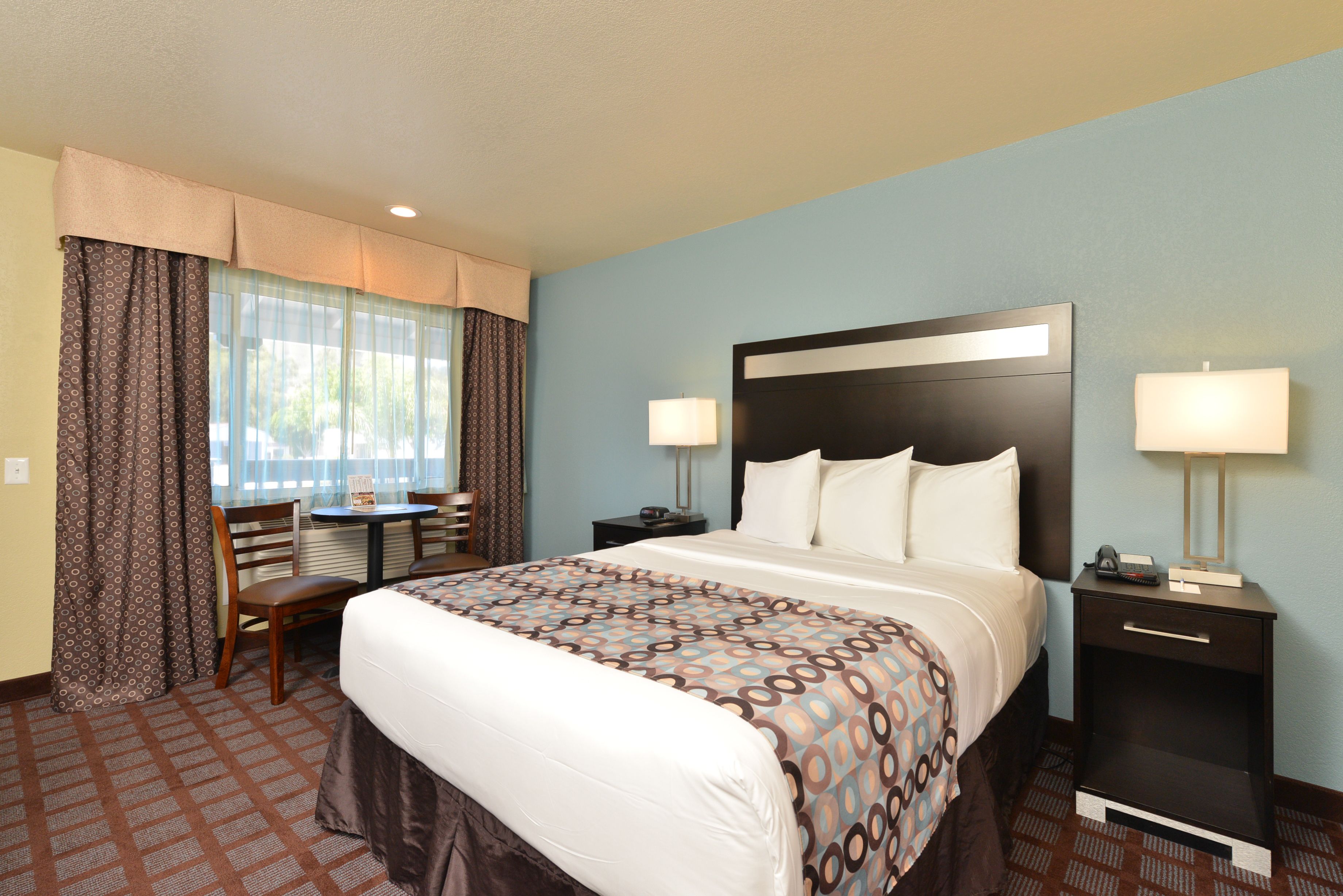 A hotel room with a large bed, two bedside tables with lamps, a desk with a chair, and a window with curtains.