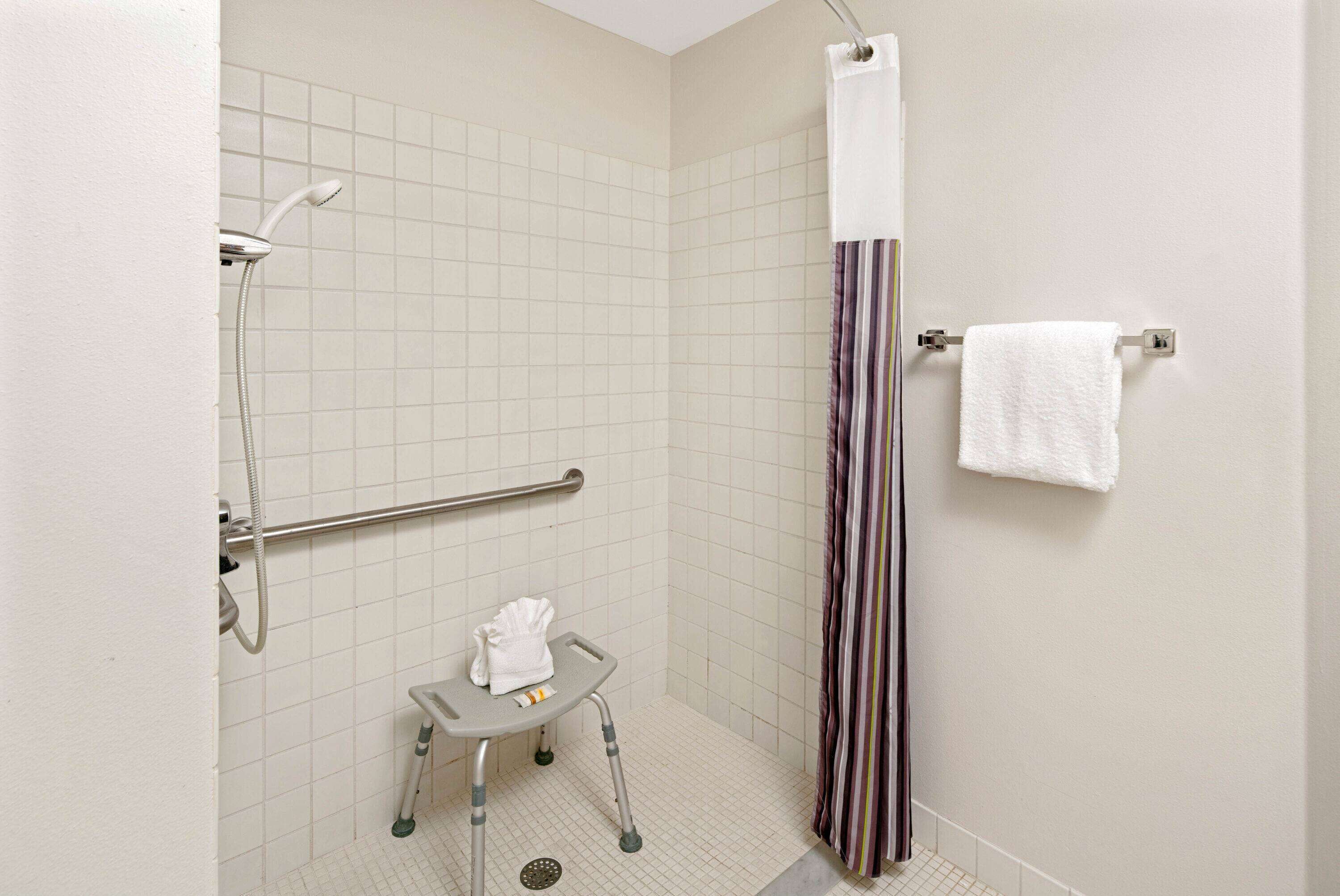 A bathroom with a shower, a grab bar, a small stool with a towel, and a striped shower curtain.