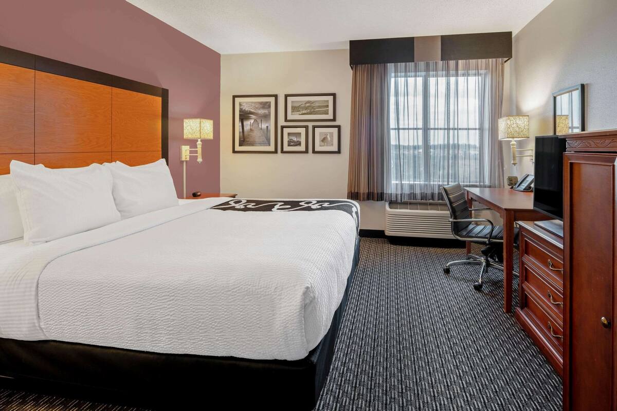 A hotel room with a large bed, a desk with a chair, a dresser, a TV, and a window with curtains.