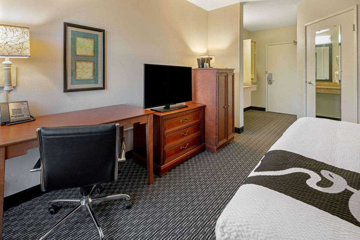 A hotel room with a bed, a desk with a chair, a television, a wardrobe, and a bathroom visible through an open door.
