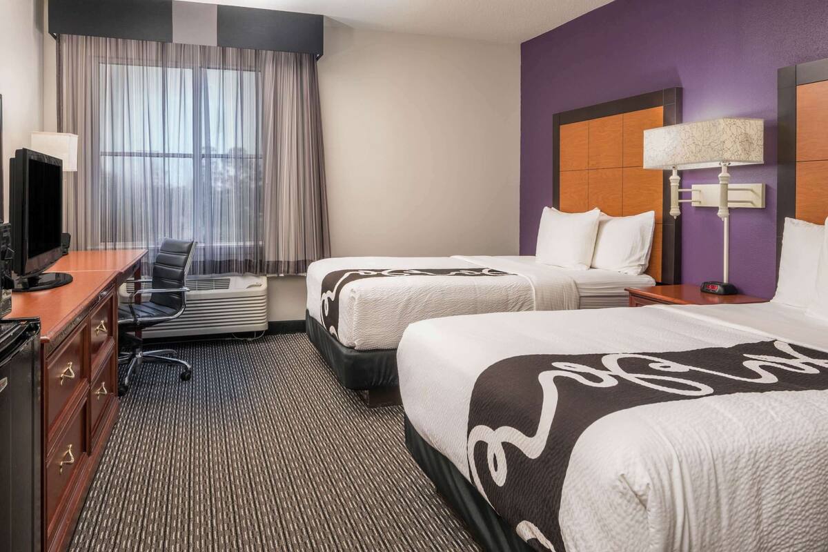 A hotel room with two beds, a desk with a TV, a chair, and a window with curtains.