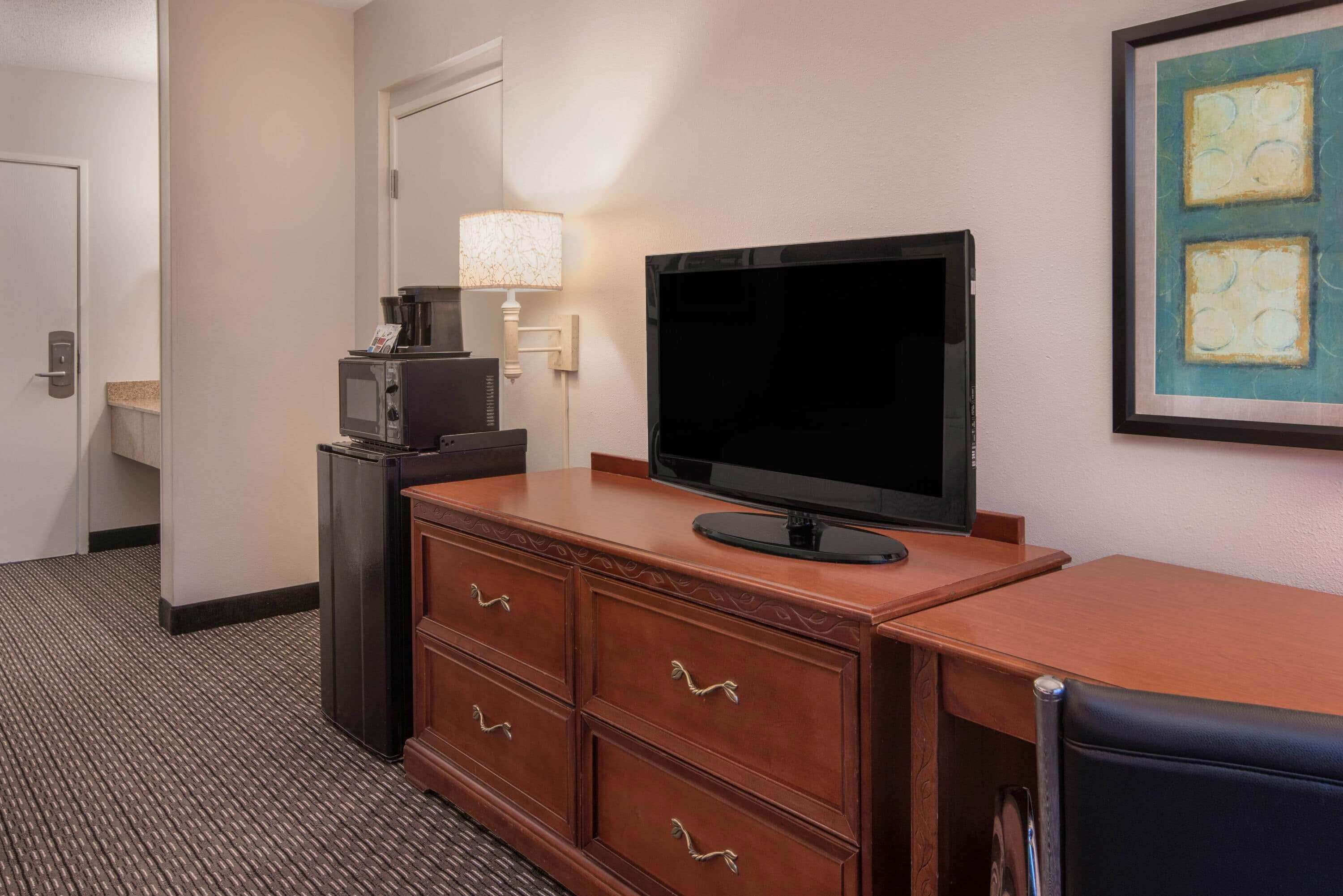 Habitación de hotel con escritorio de madera, televisión de pantalla plana, microondas, lámpara y una obra de arte enmarcada en la pared.