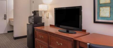 A hotel room with a wooden desk, a flat-screen TV, a microwave, a lamp, and a framed artwork on the wall.