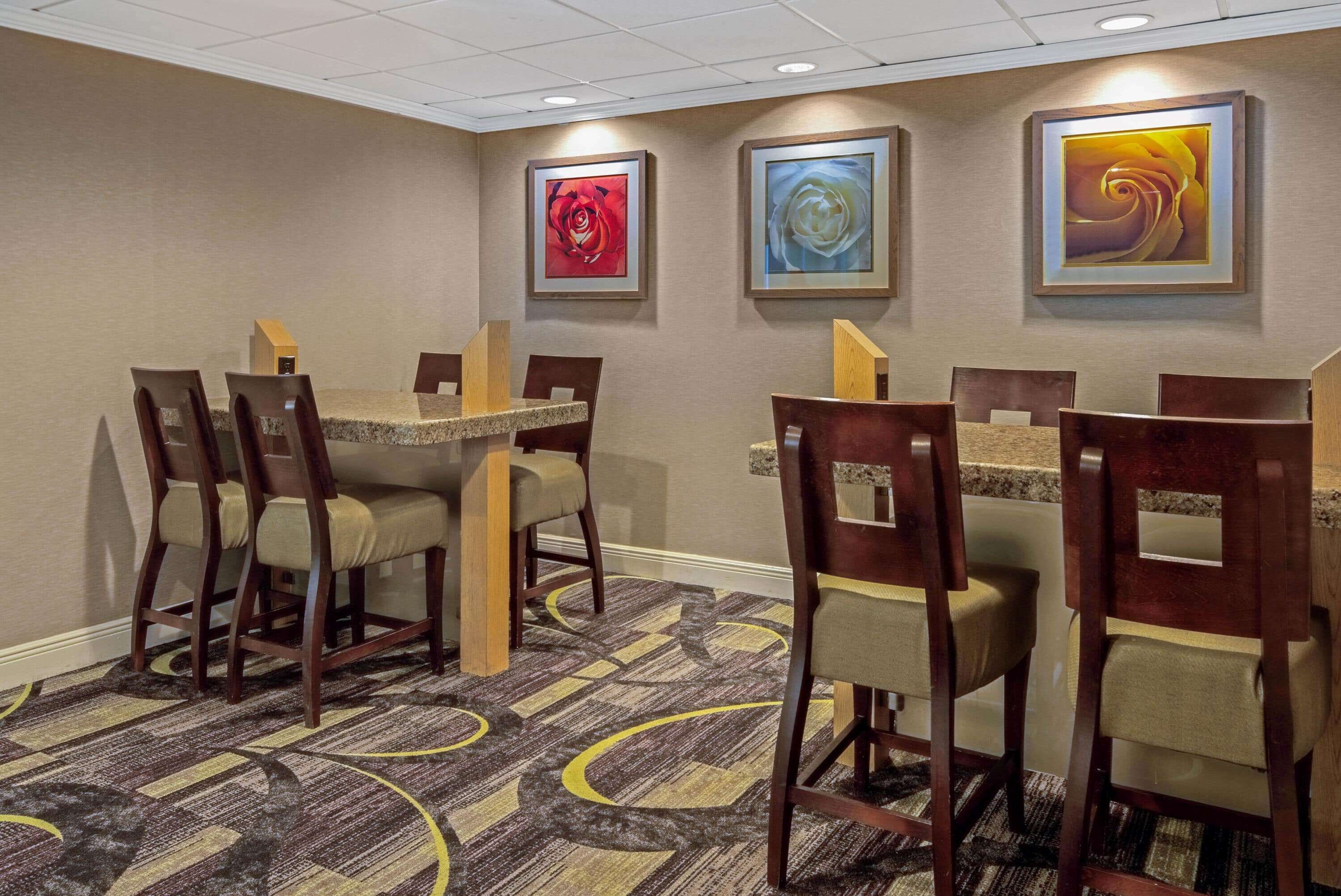 A hotel room with a dining area featuring wooden chairs and tables, a patterned carpet, and three framed artworks on the wall.