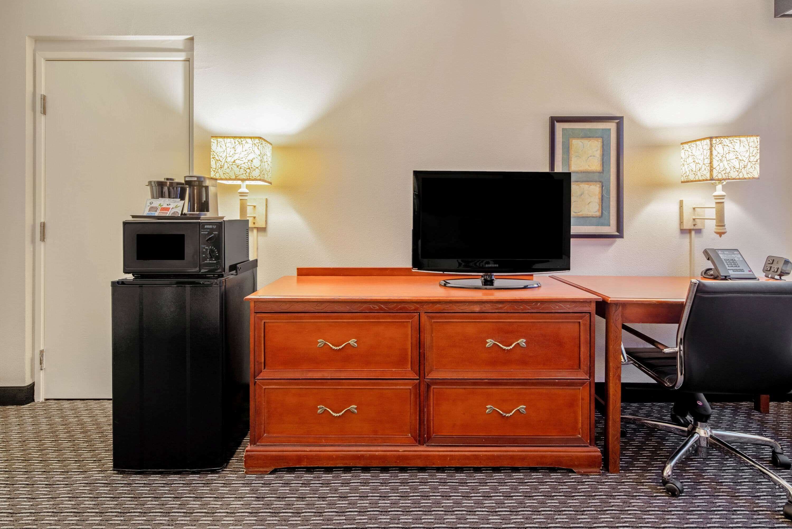 Habitación de hotel con un escritorio de madera, televisión, minibar y dos lámparas de pared.