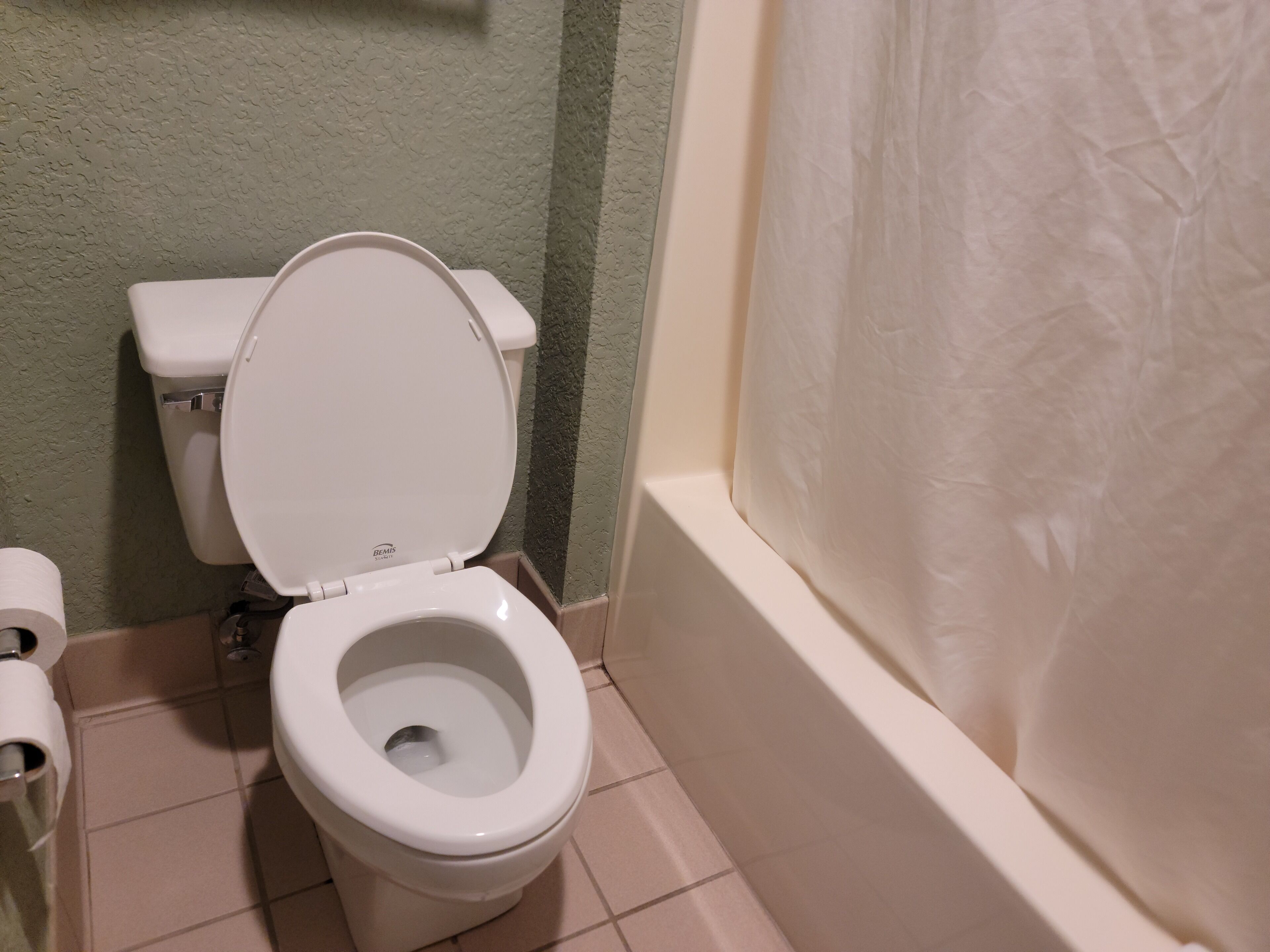 Un inodoro blanco con la tapa bajada, un rollo de papel higiénico y una cortina de ducha en un baño con paredes de textura verde y suelo de baldosas.