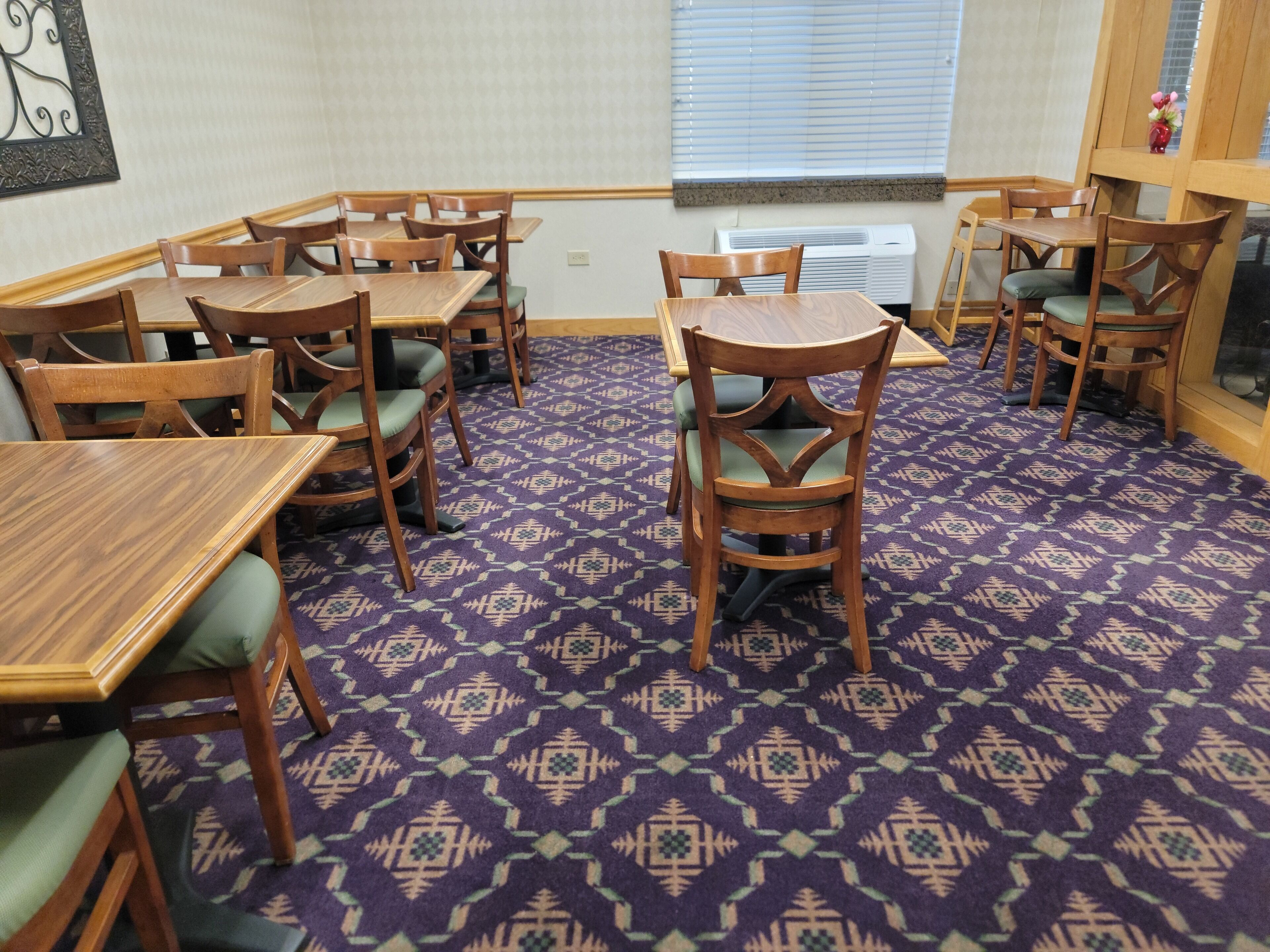 A dining area with wooden tables and chairs, patterned carpet, and a window with blinds.