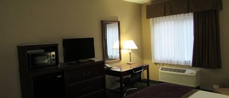 A hotel room with a bed, a desk with a lamp, a television, a microwave, and an air conditioning unit.