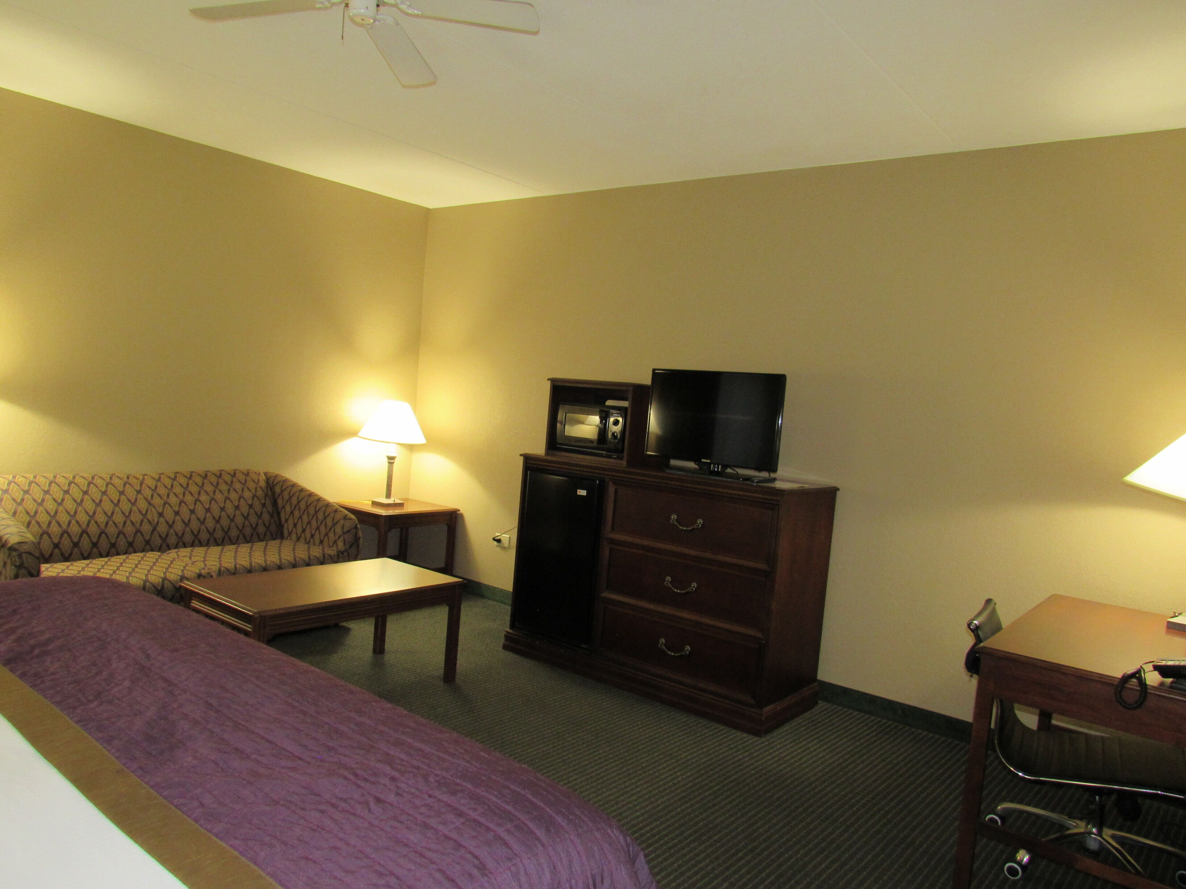 A hotel room with a bed, a sofa, a desk, a dresser, a television, and a ceiling fan.