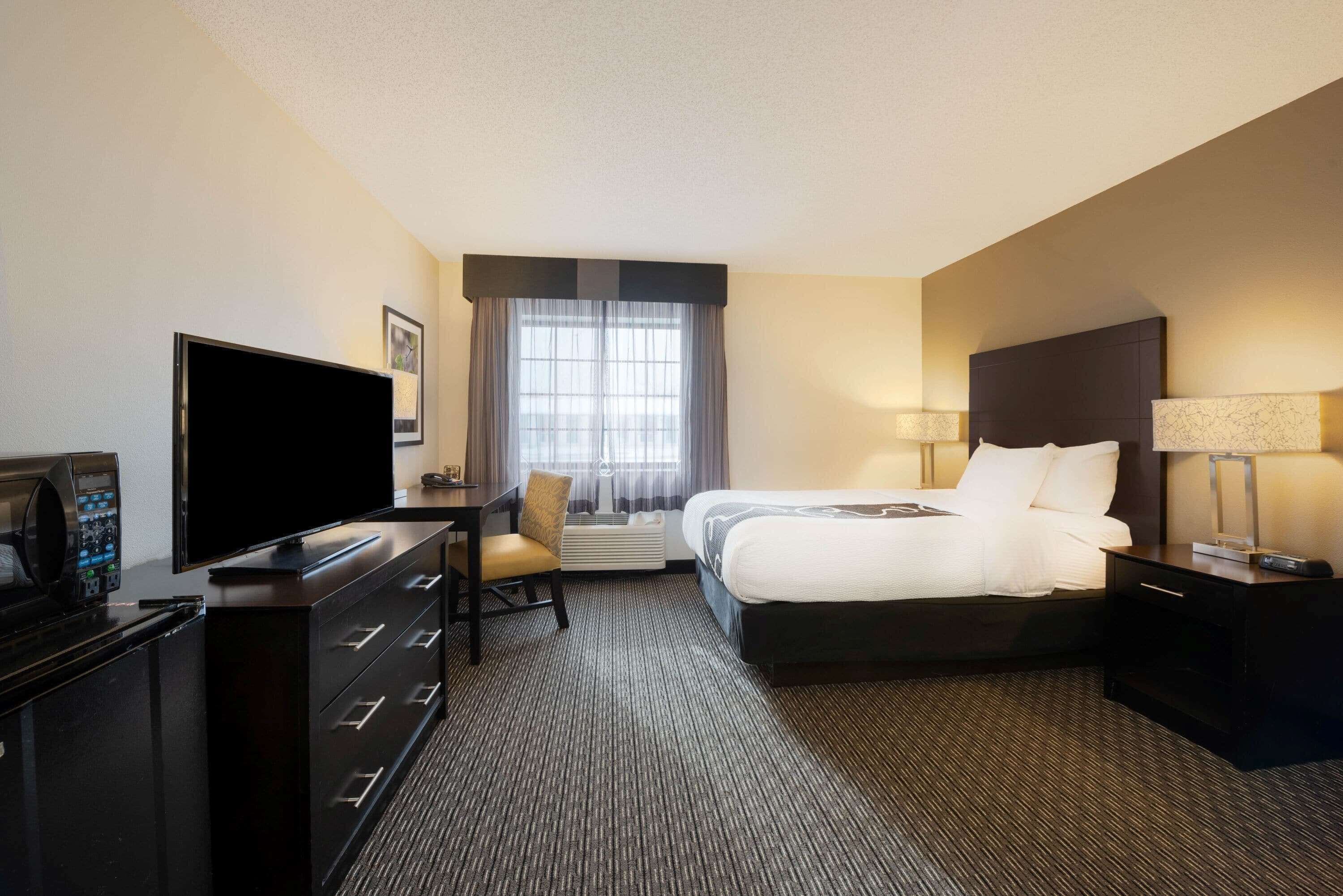 A hotel room with a bed, a desk with a flat-screen TV, a chair, a dresser, and a window with curtains.