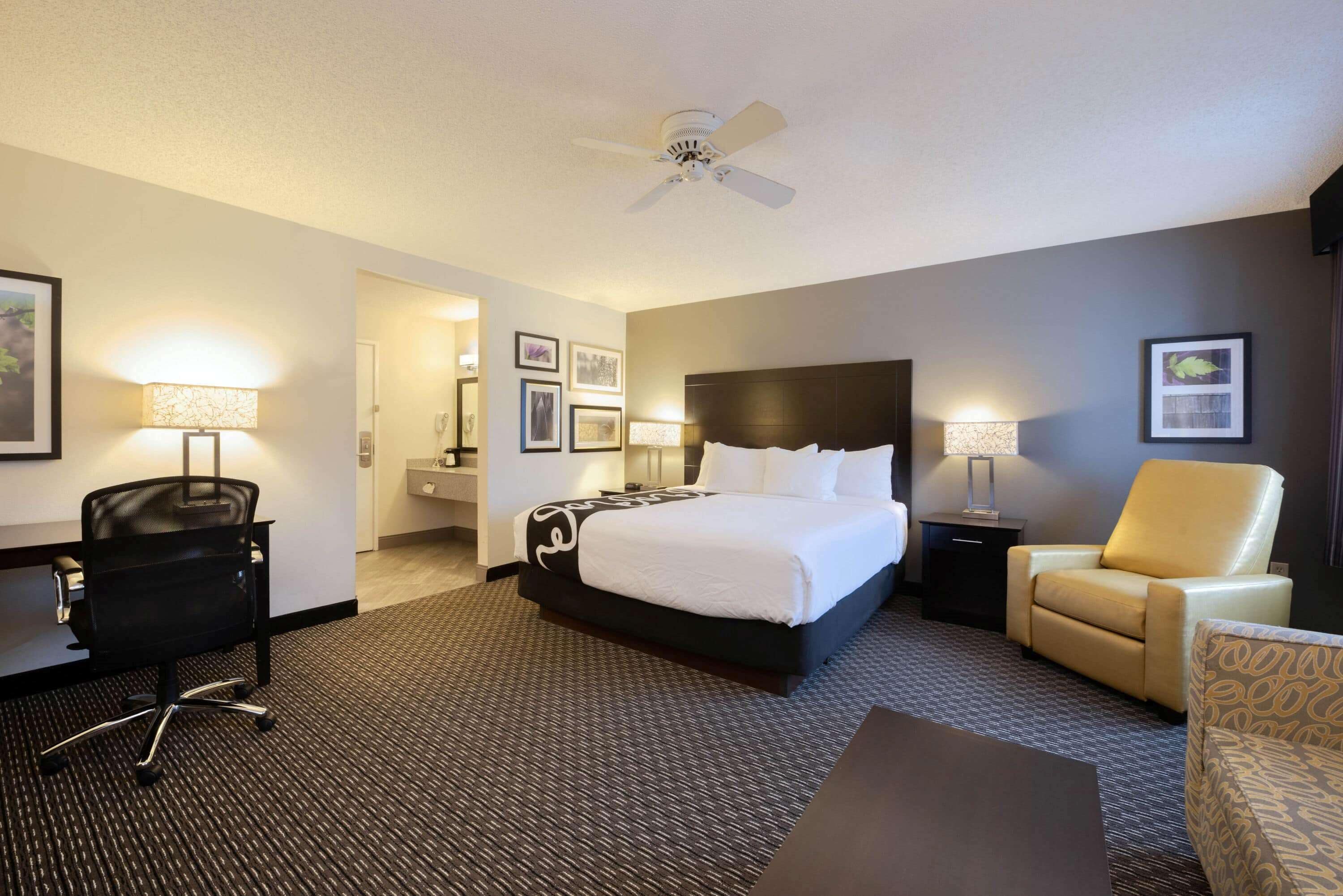 A hotel room with a large bed, a desk, a chair, a desk lamp, a standing lamp, and a ceiling fan.