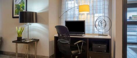 A hotel room with a desk, a computer, a lamp, a chair, a printer, and a window with curtains.