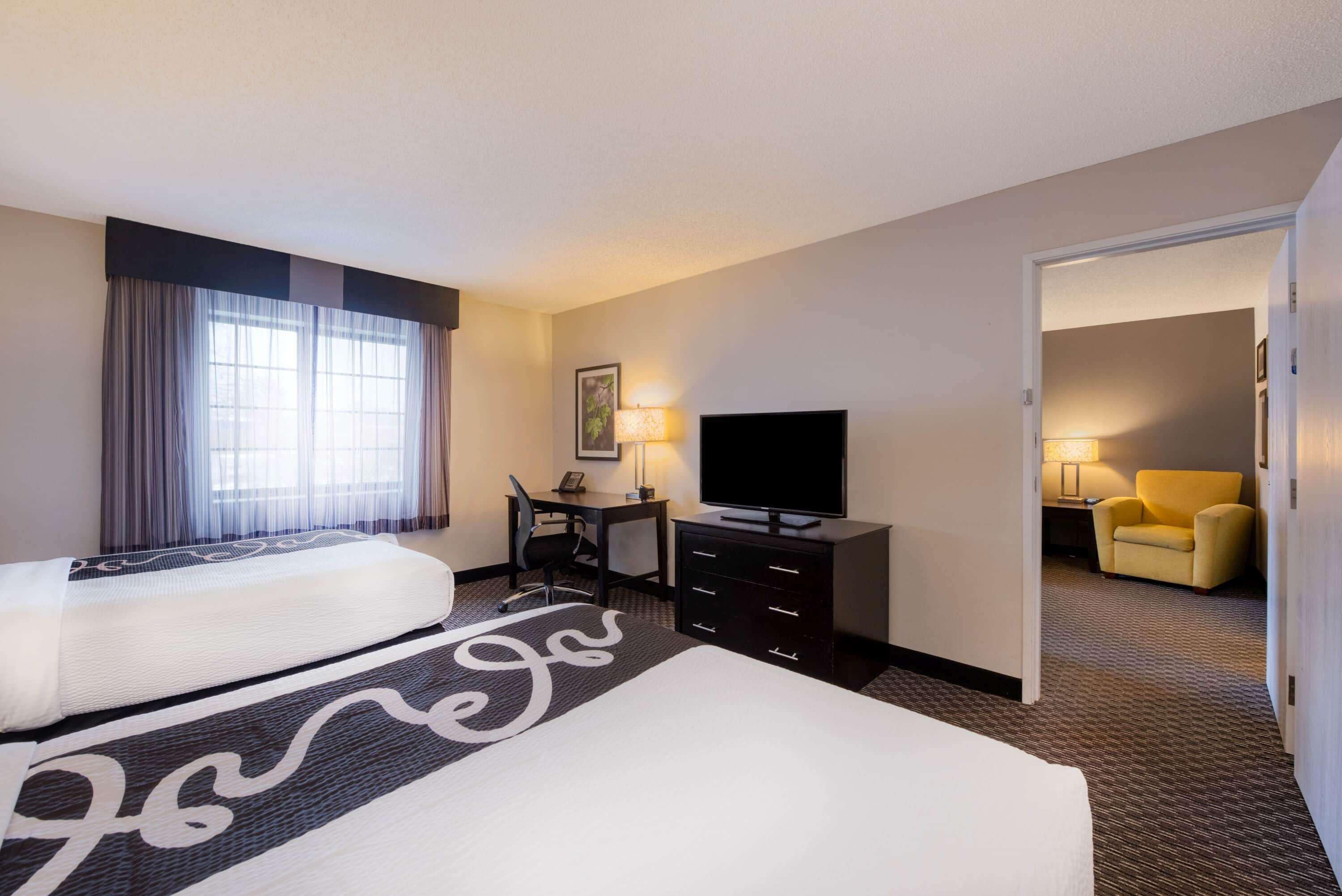 A hotel room with two beds, a desk with a chair, a television, and a yellow armchair.