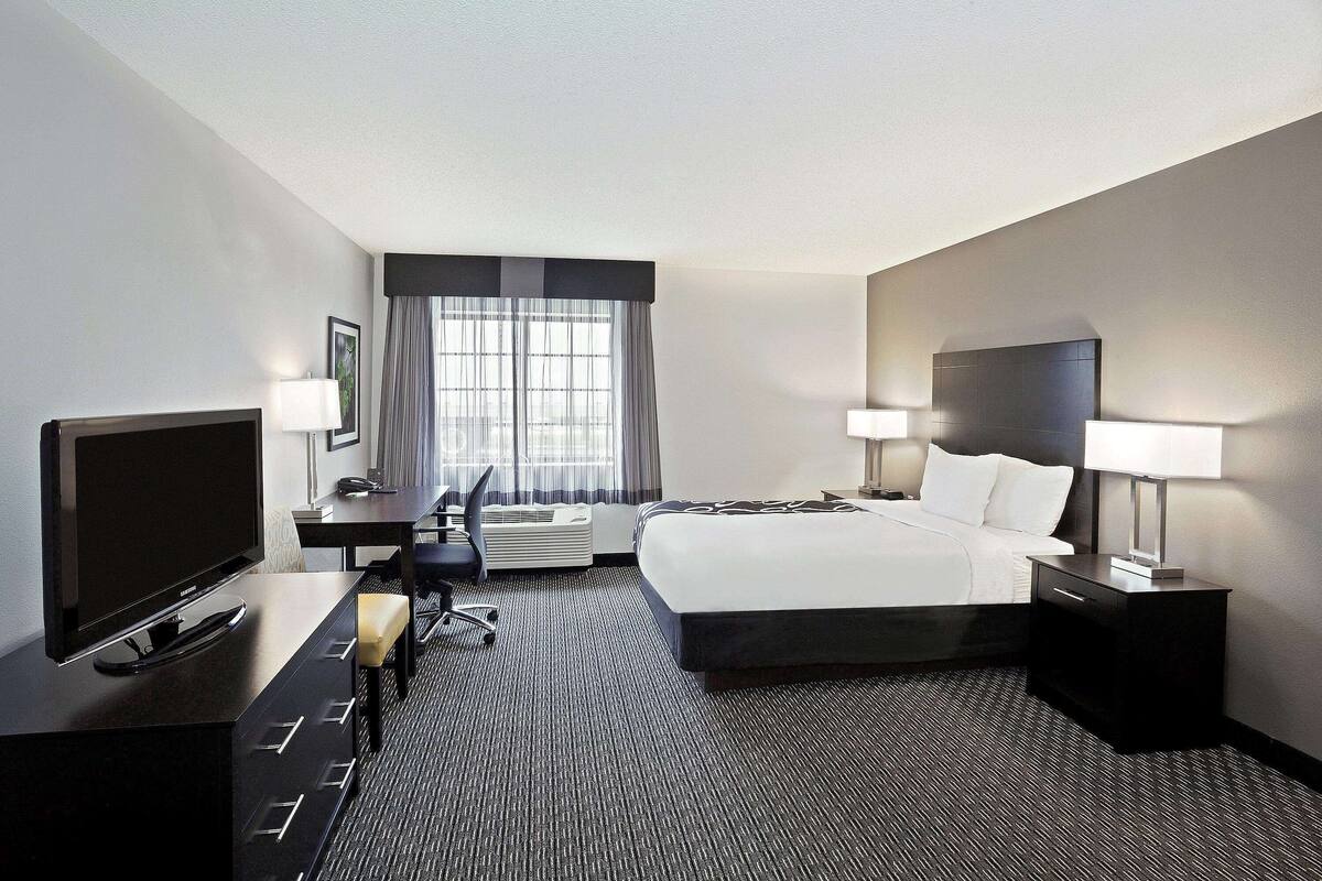 A hotel room with a large bed, a desk with a flat-screen TV, two bedside lamps, a chair, and a window with curtains.
