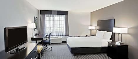 A hotel room with a large bed, a desk with a flat-screen TV, two bedside lamps, a chair, and a window with curtains.