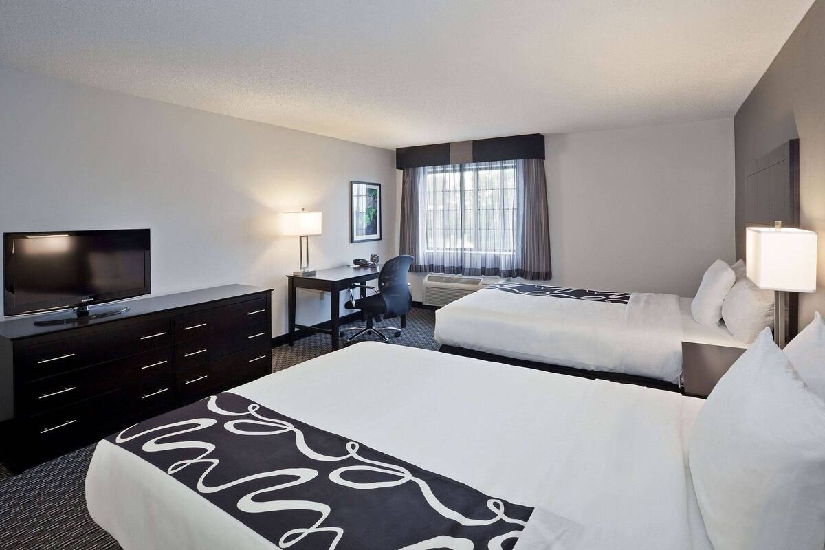A hotel room with a bed, a desk with a chair, a television, and a window with curtains.