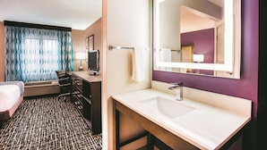 A hotel room with a bed, a desk with a computer, a bathroom with a sink and mirror, and a patterned floor. - La Quinta Inn & Suites by Wyndham Cincinnati NE - Mason (Mason)