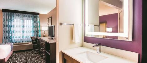 A hotel room with a bed, a desk with a computer, a bathroom with a sink and mirror, and a patterned floor.