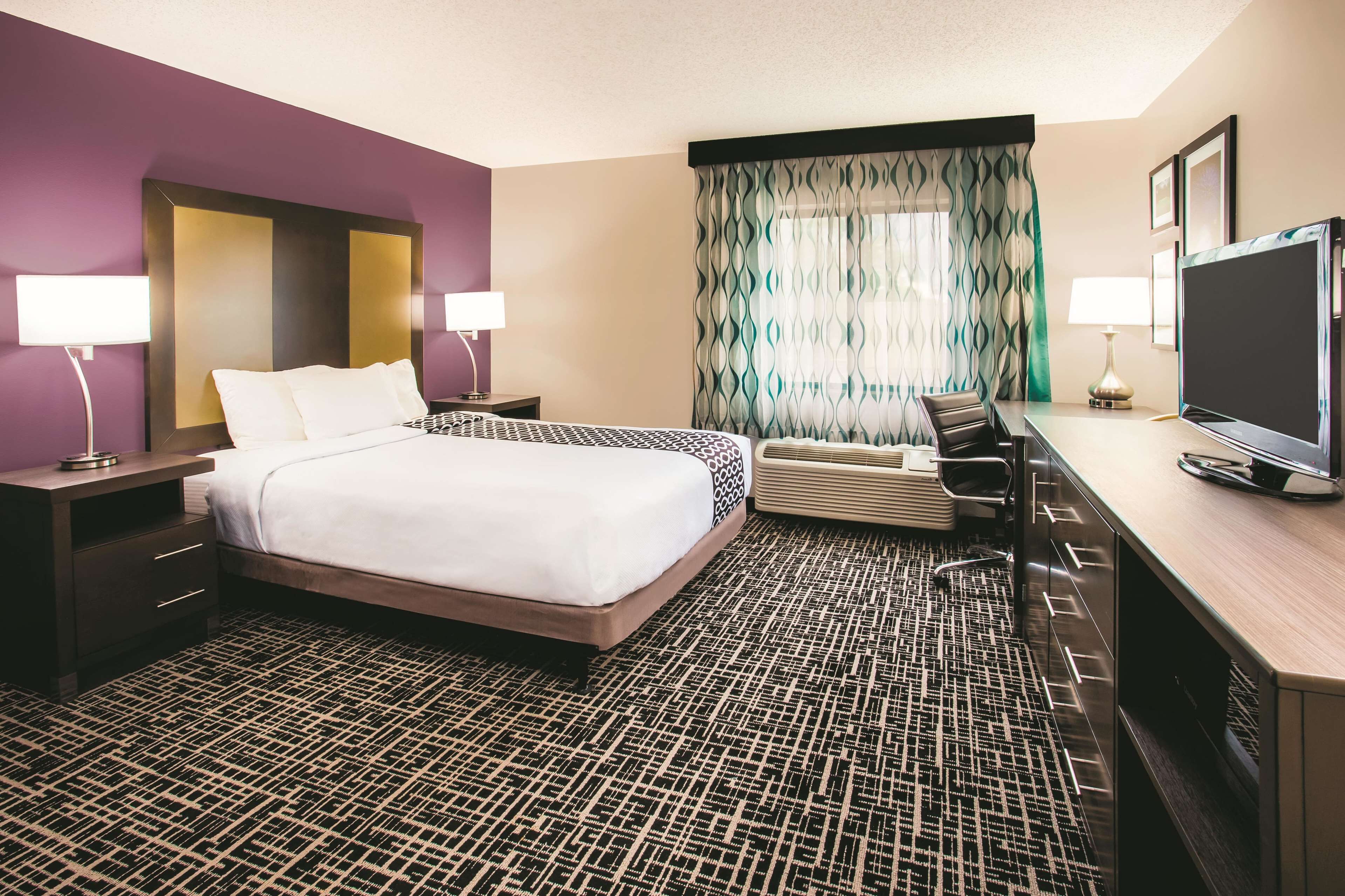 A hotel room with a large bed, a desk with a flat-screen TV, a chair, and a desk lamp.